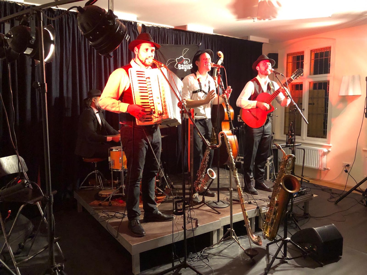Drei Musiker mit Hüten singen und spielen Akkordeon und Gitarre auf einer kleinen Bühne vor schwarzem Vorhang