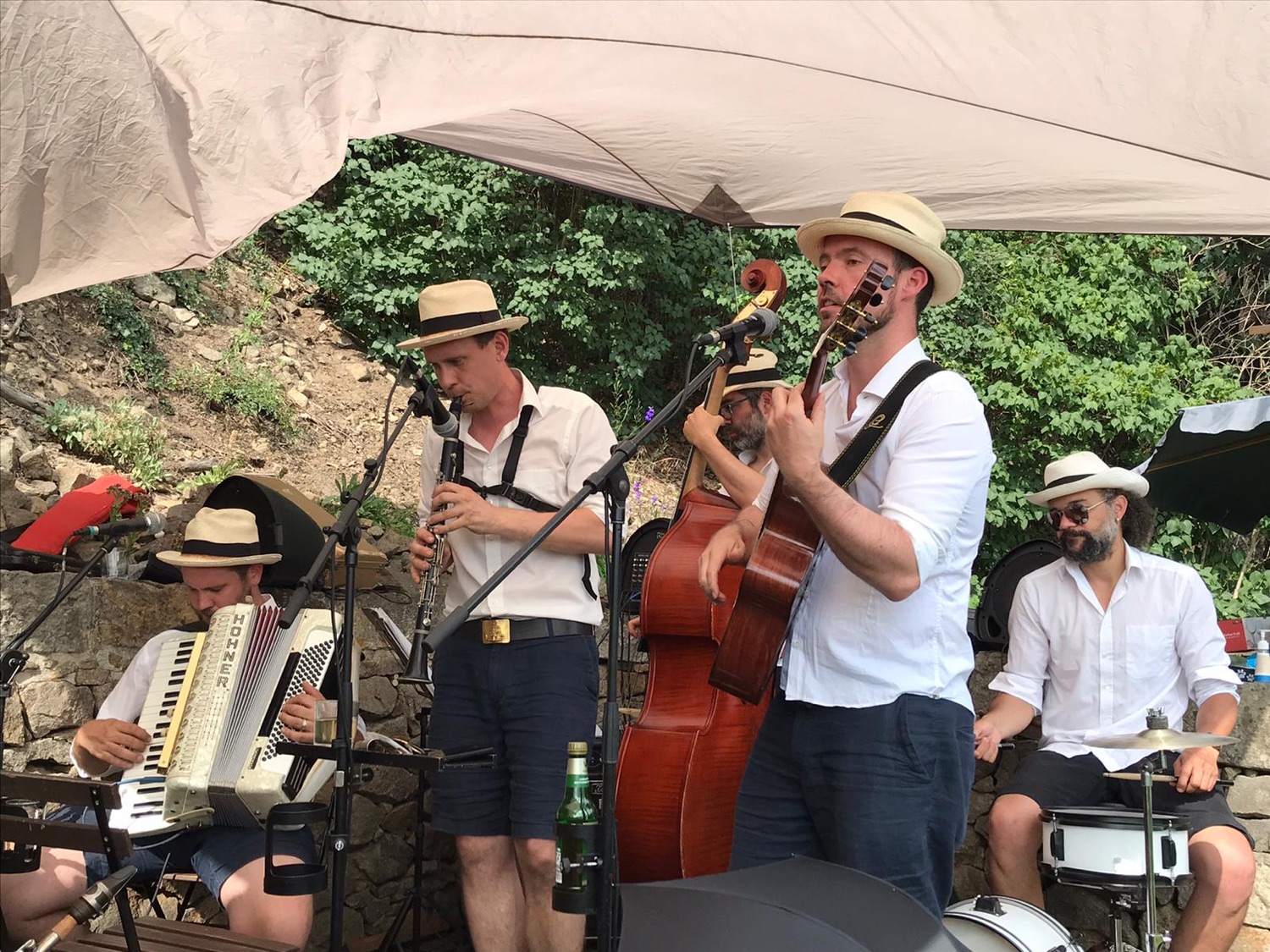 Die Band spielt unter einem weißen Sonnensegel vor grünen Pflanzen auf einer Freilichtbühne